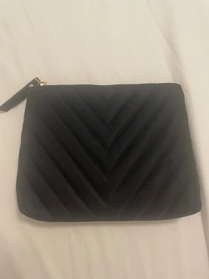 Svart quiltad Smink väska - Snygg svart smink väska  med quiltat mönster i chevronstil. Väskan har dragkedja med guldfärgad detalj och är tillverkad i ett mjukt tygmaterial. Perfekt för att bära det viktigaste när du vill ha en stilren look.