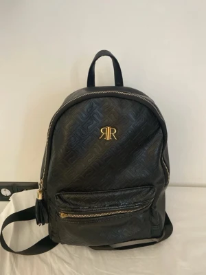 Svart ryggsäck från River Island - Snygg svart ryggsäck från River Island med guldfärgade detaljer och präglat mönster över hela väskan. Framtill finns en ficka med dragkedja och tofsdetalj. Väskan har justerbara axelband och ett praktiskt topphandtag.