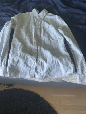 Randig skjorta från Ralph Lauren - Klassisk skjorta från Ralph Lauren med smala blå och vita ränder. Skjortan har lång ärm, knappar framtill och den ikoniska broderade loggan på bröstet. Perfekt till jeans eller chinos för en clean och stilren look.