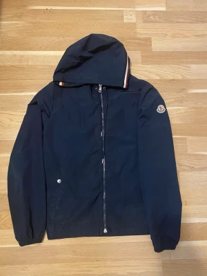 Mörkblå vindjacka från Moncler - Snygg mörkblå vindjacka från Moncler med huva och dragkedja framtill. Jackan har Moncler-logga på ärmen och diskreta rödvita detaljer på huvan. Tillverkad i ett lätt och tåligt material som passar perfekt för blåsiga dagar.