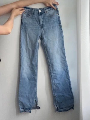 Ljusblå raka jeans med rå kant - Säljer ett par ljusblå jeans med rak passform och hög midja. Jeansen har klassiska fem fickor, knappgylf och råa, fransiga benslut för en trendig look. Tillverkade i denim, perfekta till en avslappnad stil.