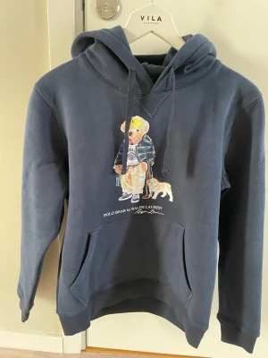 Mörkblå hoodie Polo Ralph Lauren - Snygg mörkblå hoodie från Polo Ralph Lauren med den ikoniska Polo Bear-trycket på bröstet. Hoodien har huva med dragsko och stor magficka framtill. Tillverkad i mjukt bomullsmaterial, perfekt för en chill och avslappnad stil. Storlek M kan passa S också