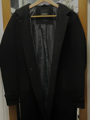 Svart ullkappa från Jack & Jones - Stilren svart ullkappa från Jack & Jones med klassisk krage och knäppning framtill. Kappan har slät insida, raka ärmar och diskreta detaljer vid ärmsluten. Passar S/M Har ett hål på insidan men går att laga 