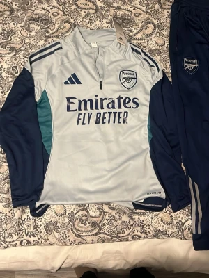 Arsenal träningströja Adidas blå - Snygg långärmad träningströja från Arsenal i ljusblått och marinblått med dragkedja vid halsen. Tröjan har klubbmärke, Adidas-logga och Emirates Fly Better-tryck på bröstet. Tillverkad i funktionsmaterial, perfekt för träning eller sport. PRIS KAN DISKUTERAS
