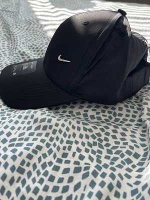 Svart Nike keps Classic99 Swoosh - Snygg svart keps från Nike, modell Classic99 Swoosh Flex. Kepsen har en böjd skärm och ett litet silverfärgat Nike-märke framtill. Tillverkad i ett mjukt och flexibelt material som sitter skönt på huvudet. Passar både tjejer och killar.