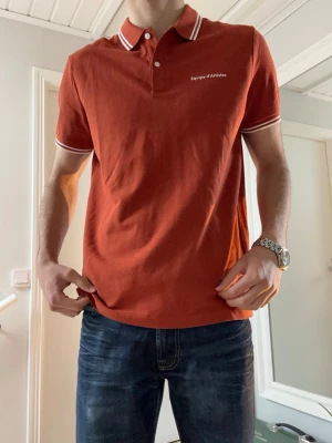 Rostbrun piké från Equipe d’Artistes - Säljer en snygg rostbrun pikétröja från H&M med vit logga på bröstet. T-shirten har korta ärmar, vit krage med ränder och knappar framtill. Tillverkad i mjuk bomull och passar perfekt till sommaren med jeans eller shorts. Priset kan diskuteras.