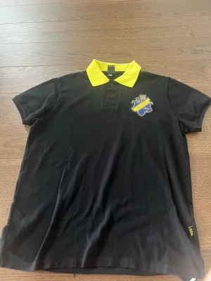 Svart AIK piké med gul krage - Svart pikétröja från AIK med gul krage och klubbmärke broderat på bröstet. Klassisk pikédesign med korta ärmar och knappar vid halsen. Perfekt för dig som vill representera AIK med stil.