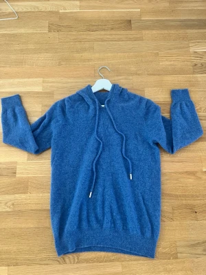Blå Kashmir hoodie med dragsko - Mysig blå hoodie med huva och långa ärmar. Tillverkad i ett mjukt, stickat material som känns skönt mot huden. Hoodien har dragsko i huvan och ribbade muddar vid ärmslut och nederkant. Perfekt för en avslappnad och chill stil. Är 184cm och väger 72kg och är lite för tajt. Har använts 2 gånger och inte mer.