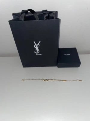 Y.S.L armband - Snyggt och stilrent armband i guldfärgad metall. Armbandet har en tunn kedja och ett Y.S.L logotyp-hänge i mitten. Perfekt accessoar för dig som gillar detaljer och klassisk design.
