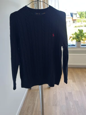 Mörkblå kabelstickad tröja Polo Ralph Lauren - Snygg mörkblå kabelstickad tröja från Polo Ralph Lauren med rund halsringning och den klassiska röda logotypen broderad på bröstet. Tröjan har långa ärmar och ribbade muddar vid ärmslut och nederkant. Perfekt för en stilren look.