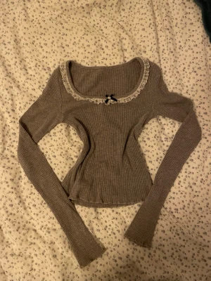Grå ribbad topp med spets och rosett - Supersöt grå ribbad topp med lång ärm, dekorerad med vit spetskant och en liten svart rosett vid urringningen. Croppad modell som sitter tight och har en feminin, vintageinspirerad vibe. Perfekt till jeans eller kjol för en trendig look.
