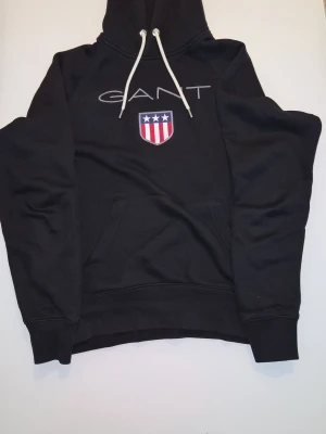 Svart hoodie från GANT med logga - Svart hoodie från GANT med stor broderad logga och sköld på bröstet. Klassisk känguruficka framtill och vit dragsko i huvan. Hoodien har ribbade muddar och är tillverkad i mjukt bomullsmaterial. Perfekt för en avslappnad och stilren look.