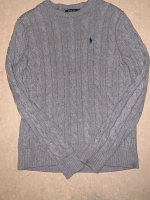 Grå kabelstickad tröja Polo Ralph Lauren - Snygg grå kabelstickad tröja från Polo Ralph Lauren med rund halsringning och den klassiska blå loggan broderad på bröstet. Tröjan har långa ärmar och ribbade muddar vid ärmslut och nederkant. Perfekt för lager på lager och stilrena outfits.