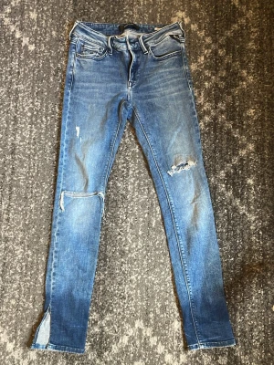  slitna skinny jeans från reaplay med slits - Säljer ett par blå skinny jeans med slitningar på knäna och slits vid bensluten. Jeansen har klassisk femficksdesign och är i stretchigt material för en tight passform. Perfekta för en avslappnad och trendig look. W28/L30 bra skick har en liten fläck på rumpan. Passningsformen är new luz