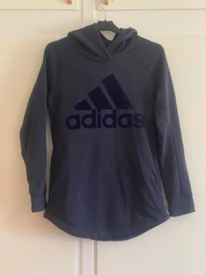 Mörkblå hoodie från Adidas - Snygg mörkblå hoodie från Adidas med stor logga framtill. Hoodien har långa ärmar, huva och en avslappnad passform. Tillverkad i mjukt material som känns skönt mot huden. Perfekt för en sportig och casual stil.