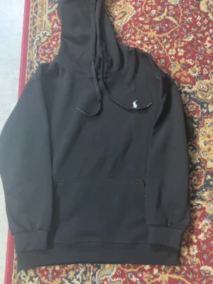 Svart hoodie från Polo Ralph Lauren - Svart hoodie från Polo Ralph Lauren med klassisk huva och dragsnören. Har en stor magficka och den ikoniska vita Polo-loggan broderad på bröstet. Tillverkad i mjukt bomullsmaterial och har ribbade muddar vid ärmslut och nederkant.