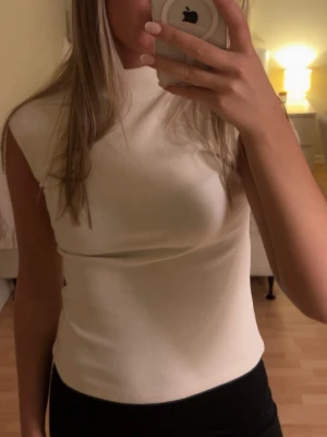 Vit ärmlös topp med hög hals - Säljer en stilren vit topp med hög hals och ärmlös design. Toppen har en tight passform och är gjord i ett mjukt material som sitter snyggt på kroppen. Perfekt att matcha med både jeans och kjol.