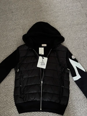 Svart pufferjacka från Moncler - Snygg svart pufferjacka från Moncler med huva och dragkedja framtill. Jackan har vadderad kropp, stickade ärmar och en vit zigzag-detalj på ena ärmen samt Moncler-logga på axeln. Perfekt för kalla vinterdagar och riktigt trendig look. Den är storlek M men passar mer som en S