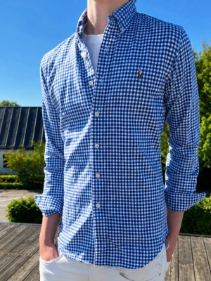 Blårutig skjorta från Ralph Lauren - Snygg blårutig skjorta från Ralph Lauren med klassisk button-down krage och långa ärmar. Skjortan har vita knappar och den ikoniska broderade loggan på bröstet. Perfekt för en stilren och avslappnad look.