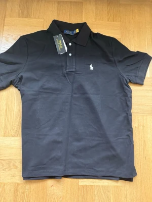 Ralph Lauren svart pikétröja – storlek M - Klassisk kortärmad pikétröja från Ralph Lauren i svart med den välkända loggan på bröstet. Ett givet basplagg i garderoben som passar till absolut allt, oavsett om det är jeans eller chinos. ✔ Storlek: M ✔ Märke: Ralph Lauren ✔ Färg: Svart ✔ Hög kvalitet i tyget med skön krage 📩 Skicka ett meddelande vid frågor eller prisförslag! 🛍 Lägg till i ett paket för att spara upp till 20% rabatt! 📦 Garanterad leverans samma eller nästa dag 🌟 