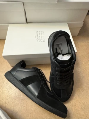  Maison Margiela Gats - Stilrena svarta sneakers från Maison Margiela med ovandel i både mocka och läder. Klassisk låg modell med svarta skosnören och diskret logga på plösen. Perfekta för dig som gillar minimalistisk och clean stil. Helt oanvända 