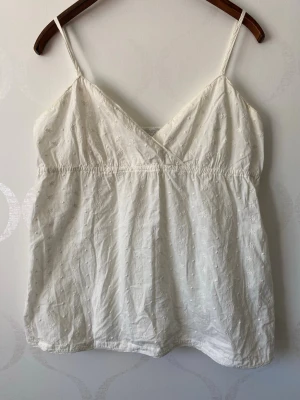 Vit broderad linnetopp med smala band - Söt vit linnetopp med tunna axelband och v-ringning. Toppen har broderade små blommor och resår under bysten för en skön passform. Perfekt till sommaren och enkel att matcha med jeans eller kjol.