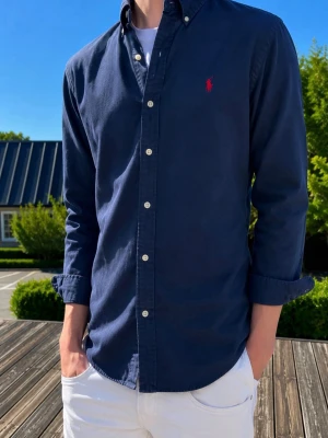 Mörkblå skjorta från Ralph Lauren - Klassisk mörkblå skjorta från Ralph Lauren med knappslå och vit knappdetalj. Skjortan har lång ärm och den ikoniska röda logotypen broderad på bröstet. Perfekt till jeans eller chinos för en stilren look.