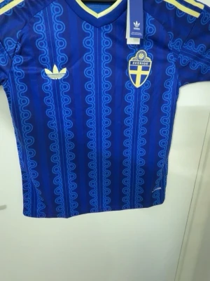Blå Sverige fotbollströja Adidas - Snygg blå fotbollströja från Adidas med Sveriges landslagsemblem på bröstet och retroinspirerat mönster i olika blå nyanser. Tröjan har gula detaljer på axlarna och runt halsen samt Adidas-logga på bröstet. Perfekt för match eller träning.