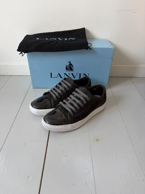 Svarta Lanvins - Snygga svarta sneakers från Lanvin med glansig lackyta och vita sulor.  Box och dustbag. Skorna har grå skosnören och klassisk låg profil. Perfekta för dig som vill ha en stilren och exklusiv look. Skorna är använt skick och har tecken på användning, se följande bilder. Orginalsula ingår inte. Det är bara att skriva vid frågor! Mvh Viggo