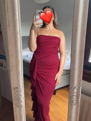 Vinröd axelbandslös långklänning - Säljer en vinröd axelbandslös långklänning med draperade detaljer och figurnära passform. Klänningen har en snygg volangdetalj på sidan och är perfekt för speciella tillfällen. Materialet känns mjukt och följsamt mot kroppen.