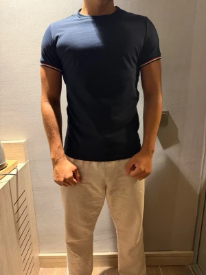 Moncler tshirt - Snygg marinblå t-shirt från Moncler med klassisk rund hals och korta ärmar. Ärmsluten har rödvita ränder och Moncler-logga sitter på vänster ärm. Tillverkad i mjuk bomull för skön passform och stilren look.