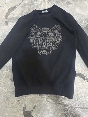 Svart Kenzo sweatshirt  - Svart sweatshirt från Kenzo med det ikoniska tigermotivet broderat på bröstet och Kenzo-loggan i mitten. Tröjan har rund halsringning, långa ärmar och ribbade muddar vid ärmslut och nederkant. Perfekt för en trendig streetwear-look.