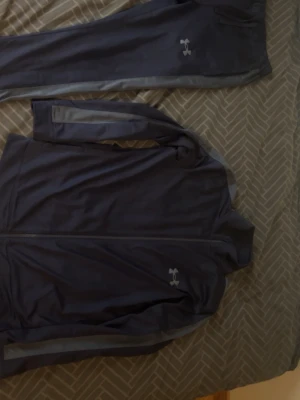 Mörkblå träningsset från Under Armour - Säljer ett mörkblått träningsset från Under Armour med matchande jacka och byxor. Setet har dragkedja på jackan och diskreta loggor på både jacka och byxa. Materialet är mjukt och stretchigt, perfekt för träning eller uppvärmning.