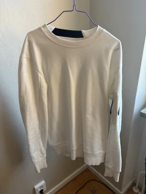 Vit sweatshirt från C.P. Company - Säljer en stilren vit sweatshirt från C.P. Company med deras ikoniska svarta linsdetalj på ärmen. Tröjan har rund halsringning, ribbade muddar och är gjord i mjukt bomullsmaterial. Perfekt för dig som gillar minimalistisk och modern streetwear.