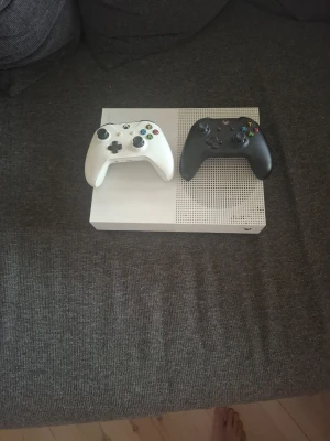 Xbox One S med två handkontroller - Säljer en vit Xbox One S med två trådlösa handkontroller, en vit och en svart. Konsolen har en stilren, rektangulär design med ventilerade detaljer på ovansidan. Perfekt för gaming och streaming. Materialet är plast och konsolen har klassisk Xbox-look.