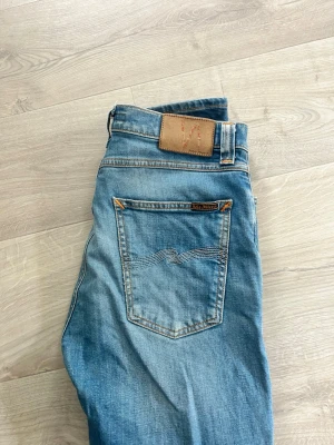 Blå jeans från Nudie Jeans - Säljer ett par klassiska blå jeans från Nudie Jeans. Passform lean dean och snygga slitningar. Jeansen har fem fickor, kontrastsömmar och en brun läderpatch med logga bak i midjan. Tillverkade i bomull för en skön och avslappnad look.