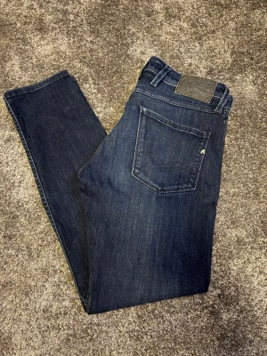 Mörkblå Replay Anbass jeans 30/30 - Snygga mörkblå jeans från Replay, modell Anbass. Klassisk femficksdesign med raka ben och diskreta sömmar. Jeansen har en stilren look och är tillverkade i mjukt denimtyg som ger en bekväm passform. Perfekta för dig som gillar en tidlös och clean stil.