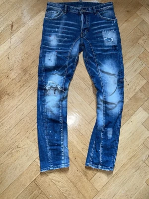 Blå distressed jeans från Dsquared2 - Säljer ett par blå jeans från Dsquared2 med slitna detaljer, färgstänk och tydliga slitningar på benen. Jeansen har en skinny fit och klassisk femficksmodell. Materialet är denim med stretch för extra komfort. Perfekt för dig som gillar en edgy och trendig stil.