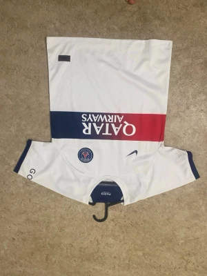 PSG matchtröja vit Nike - Snygg vit PSG matchtröja från Nike med blå och röd rand över bröstet och Qatar Airways-tryck. PSG-logga och Nike swoosh på framsidan, samt detaljer i blått och rött på ärmar och rygg. Perfekt för dig som älskar fotboll och vill representera Paris Saint-Germain.