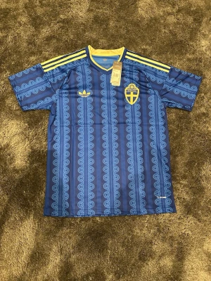 Blå Sverige fotbollströja - Snygg blå fotbollströja från Adidas med Sveriges landslagsemblem på bröstet och gula detaljer. Tröjan har ett unikt mönster i ljusblått, gula ränder på axlarna och rund halsringning med gul kant. Tillverkad i lätt och funktionellt material.