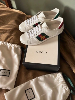 Gucci ace bee ÄKTA - Ett par snygga äkta Gucci Ace vita sneakers i läder med gröna och röda detaljer på hälen, klassiska gröna och röda sidoremsor samt broderad guldfärgad bi på sidan. Skorna har vita snören och låg profil. Kommer med originalkartong och dustbags.