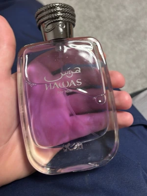 Hawas For Him parfym - Säljer en Hawas For Him parfym med en stilren, transparent glasflaska och lila vätska. Flaskan har ett dekorativt, silverfärgat lock med mönster och arabiska bokstäver på framsidan. Perfekt för dig som gillar unika dofter.