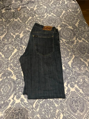 Mörkblå Lee jeans med rak passform - Säljer ett par mörkblå jeans från Lee med klassisk rak passform och fem fickor. Jeansen har Lee-lappen bak i midjan och är sydda i slitstark denim. Perfekta för dig som gillar tidlös stil och vill ha ett par jeans som funkar till det mesta. W30L32