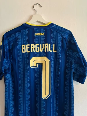 Blå Sverige matchtröja Adidas - Säljer en blå Sverige matchtröja från Adidas med gula detaljer och trycket 'Bergvall' samt nummer 7 på ryggen. Tröjan har gula ränder på axlarna, FIFA-märke på ärmen och ett unikt mönster i tyget. Perfekt för fotbollsfans!