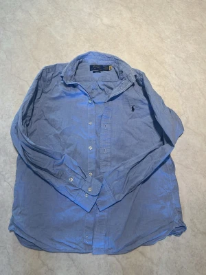 Blå skjorta från Polo Ralph Lauren - Klassisk blå skjorta från Polo Ralph Lauren med button-down krage och broderad logga på bröstet. Skjortan har långa ärmar, vita knappar och är tillverkad i bomull. Perfekt för en stilren och avslappnad look.