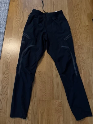 Svarta träningsbyxor Under Armour - Snygga svarta mjukisbyxor från Under Armour med smal passform och dragkedjefickor på sidorna. Byxorna har reflexdetaljer längs benen och justerbar midja med snörning. Perfekta för träning eller chill. Materialet är lätt och stretchigt.