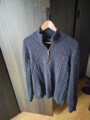 Mörkblå kabelstickad tröja Ralph Lauren - Mörkblå kabelstickad tröja från Polo Ralph Lauren med halv dragkedja och hög krage. Tröjan har ett rött broderat logomärke på bröstet, ribbstickade muddar och nederkant. Perfekt för kyliga dagar och klassisk stil.