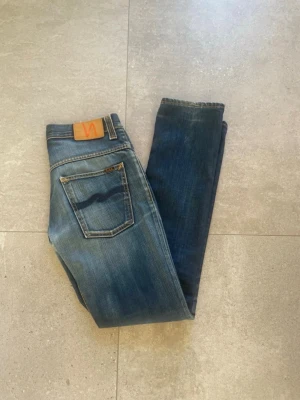 Blå jeans från Nudie Jeans - Säljer ett par klassiska blå jeans från Nudie Jeans med snygg tvätt och kontrastsömmar. Jeansen har raka ben, fem fickor och en läderpatch med logga bak i midjan. Perfekta för en avslappnad och trendig stil. Storlek W31 L34.  Längd:105cm, Midja 40cm. Tveka inte, skruv vid funderingar!!!
