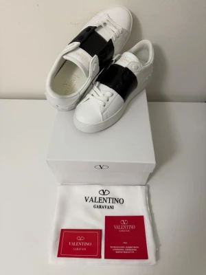 Valentino Garavani vita sneakers - Stilrena vita sneakers från Valentino Garavani med bred svart läderdetalj över vristen. Klassisk låg modell med rund tå, vita snören och präglad logga på sulan. Tillverkade i läder för en lyxig känsla och look.