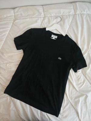 Svart t-shirt från Lacoste - Nyskick Klassisk svart t-shirt från Lacoste med den ikoniska krokodilloggan broderad på bröstet. T-shirten har rund halsringning och korta ärmar. Tillverkad i mjuk bomull för en skön och avslappnad stil.